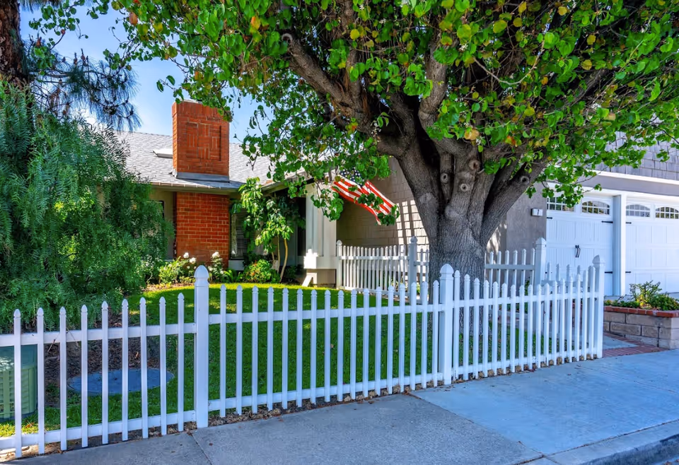 Front yard of a house with a white picket fence, large tree, and American flag.