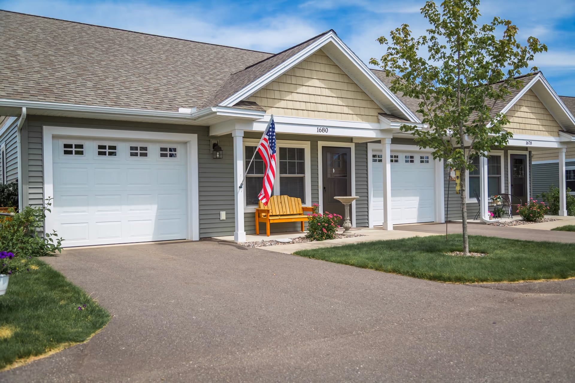Exterior view of two adjacent senior living community units with beige siding and white garage doors. Each unit has a small porch area with seating and decorative plants. One porch features an American flag and a wooden bench, while the other has a small table and chairs. A young tree is planted in the grassy area between the driveways.