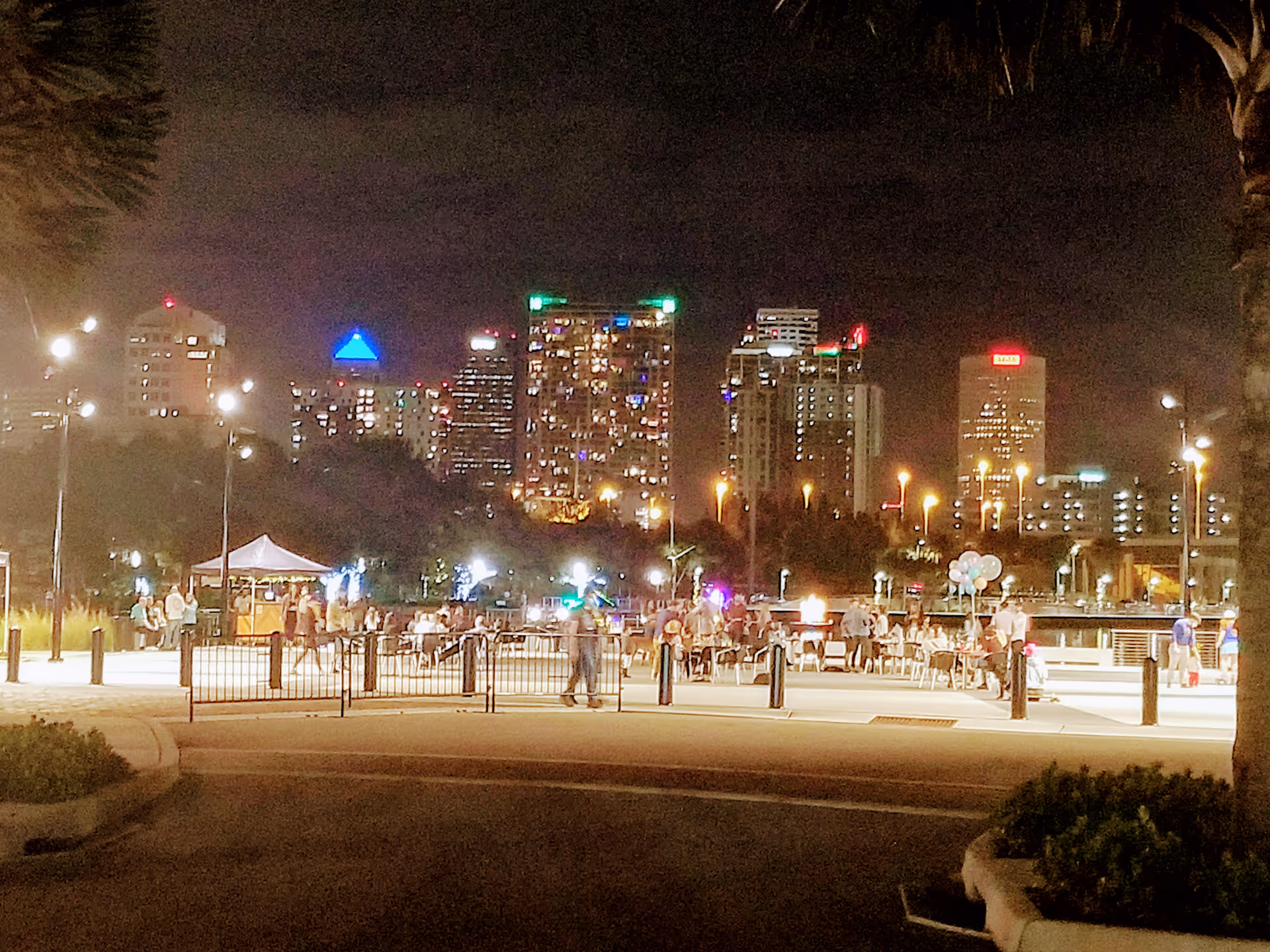 Nighttime waterfront promenade with people gathering near tents against a lit city skyline.