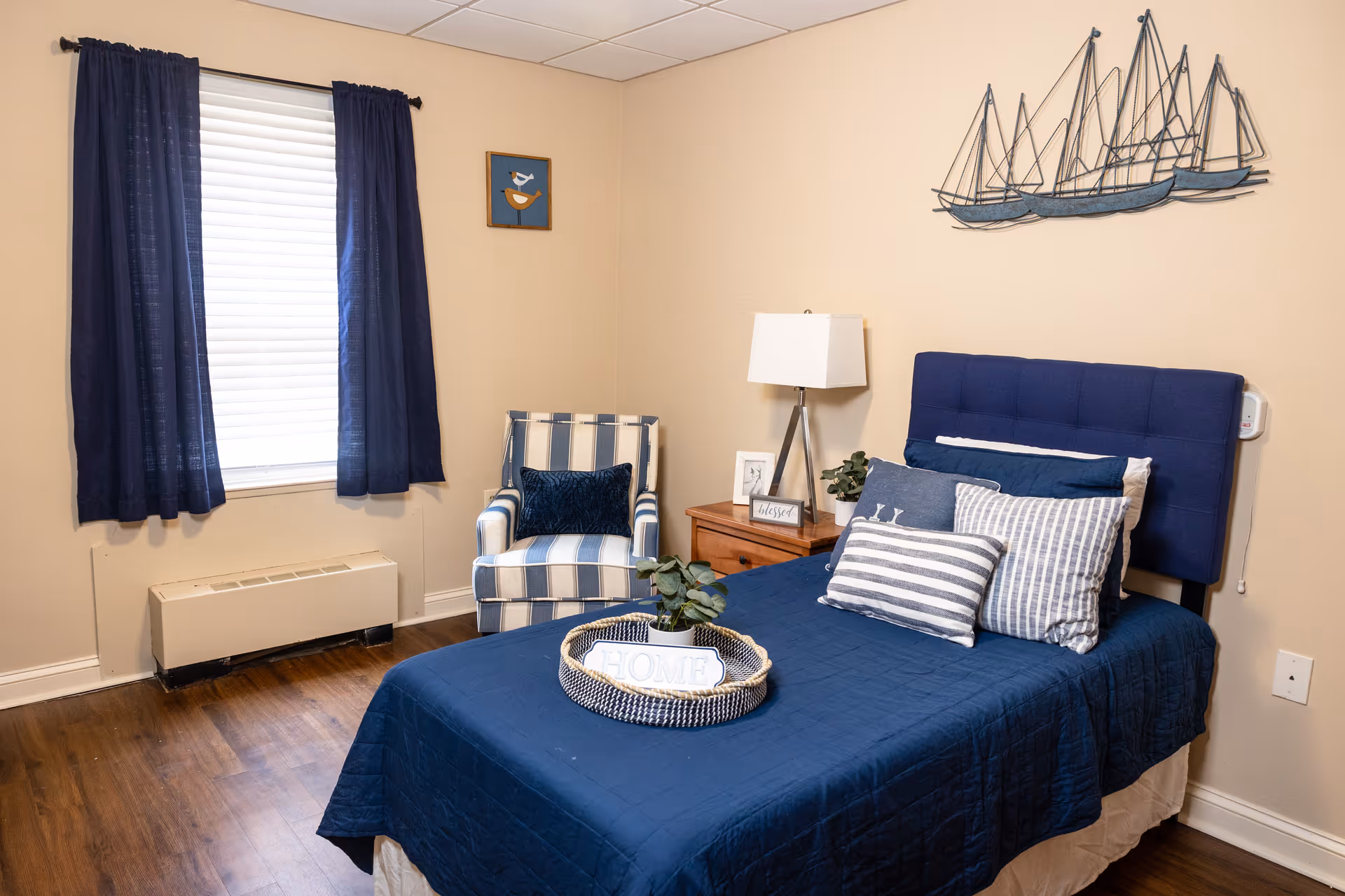 Cozy bedroom with navy bedding, blue curtains, striped chair, and sailboat wall art.