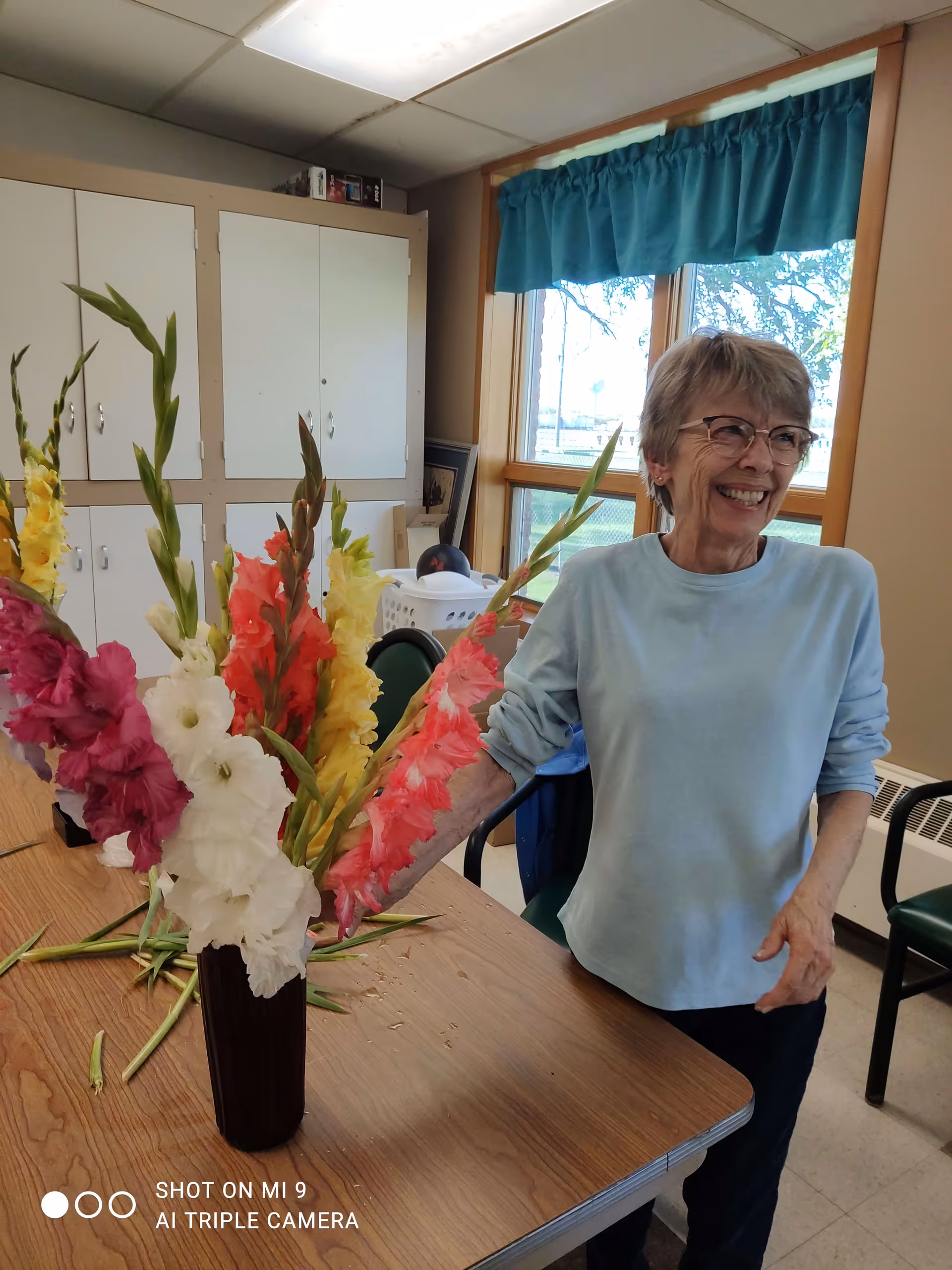 An elderly woman with short gray hair and glasses, wearing a light blue long-sleeve shirt, stands smiling next to a wooden table with a vase of colorful gladiolus flowers. The room has large windows with teal curtains, white cabinets, and a laundry basket in the background.