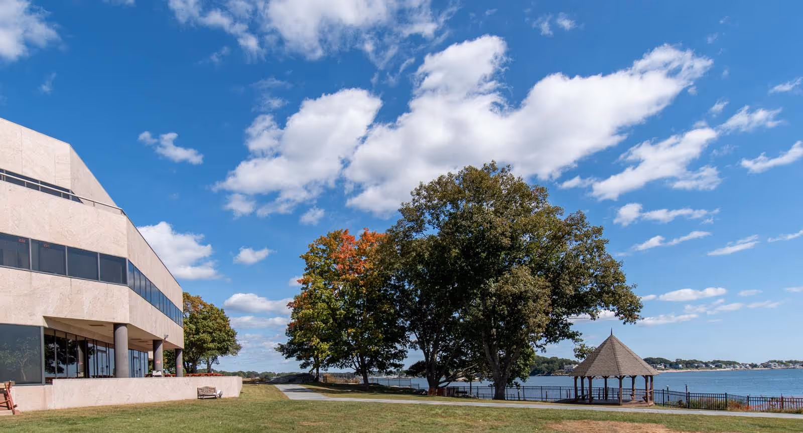 Exterior view of a beige building with a grassy lawn, trees, a gazebo, and a waterfront.
