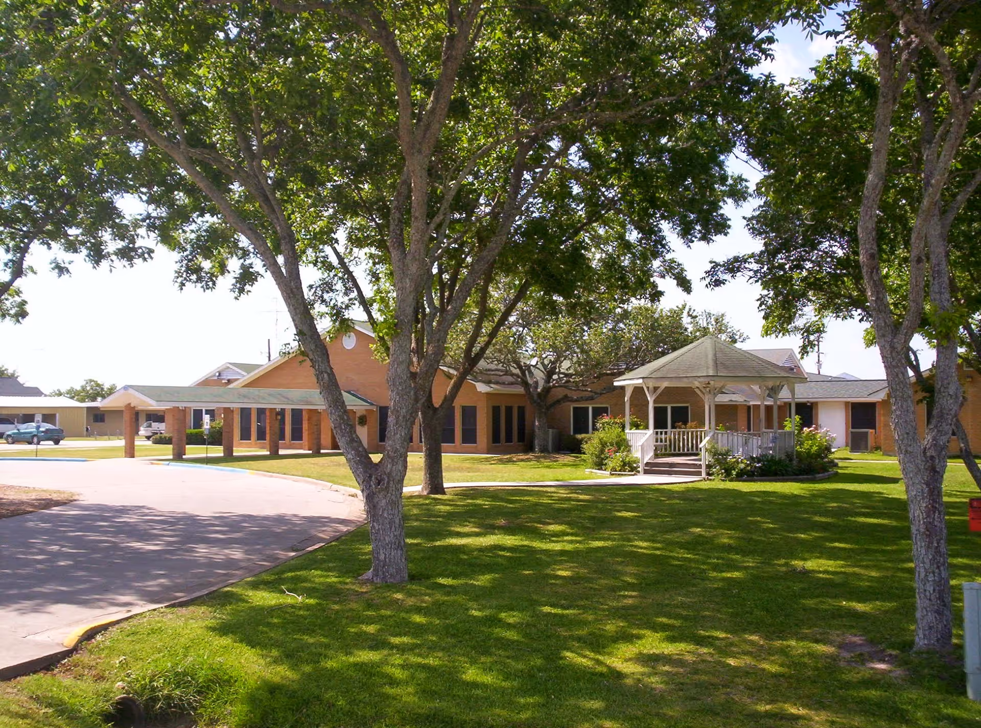 View of a single-story brick building with a green lawn and several large trees in the foreground. There is a white gazebo on the lawn near the building, and a paved driveway curves around the grass area.