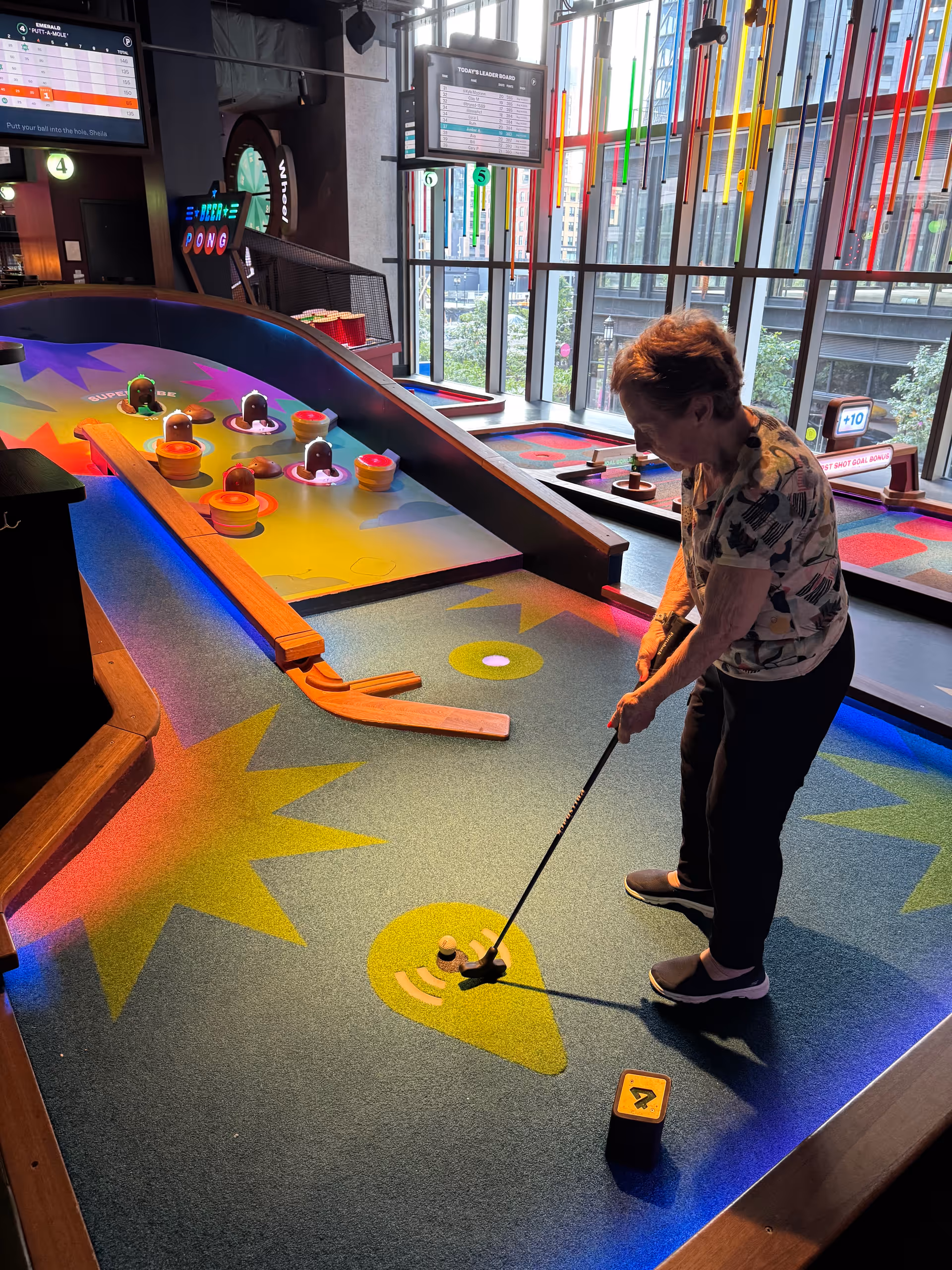 A woman putting on a brightly lit indoor mini-golf course with colorful obstacles and large windows in the background.