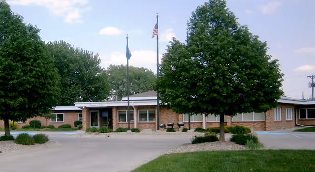 Brick senior living facility exterior with flagpoles, trees, and a paved entrance.