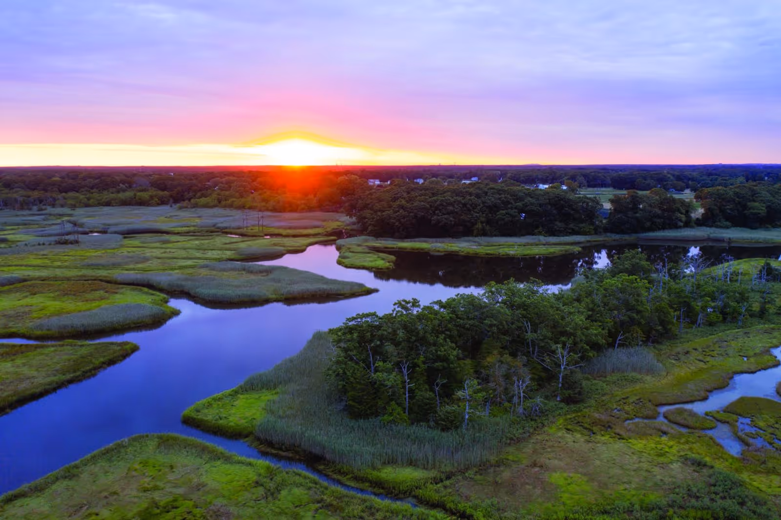 Aerial view of marshy wetlands and winding waterways at sunset under a colorful sky.