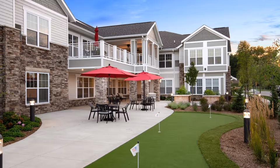 Outdoor courtyard of a senior living building with patio tables under red umbrellas and a small putting green.