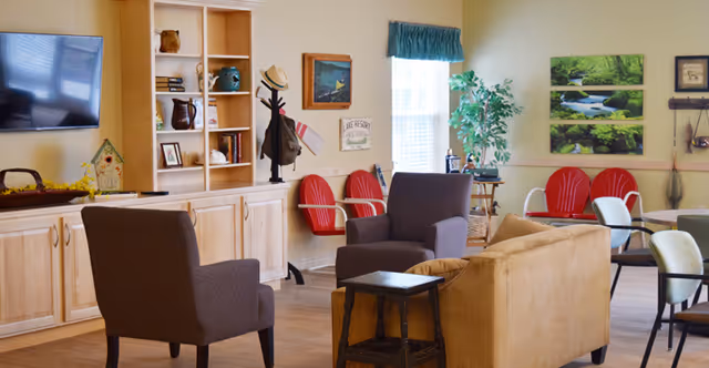 Bright communal living room with sofas and armchairs arranged around tables, a wall-mounted TV, shelving, and decorative artwork.