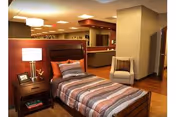 A furnished bedroom in a senior facility with a wooden bed, nightstand, lamp, and striped bedding.