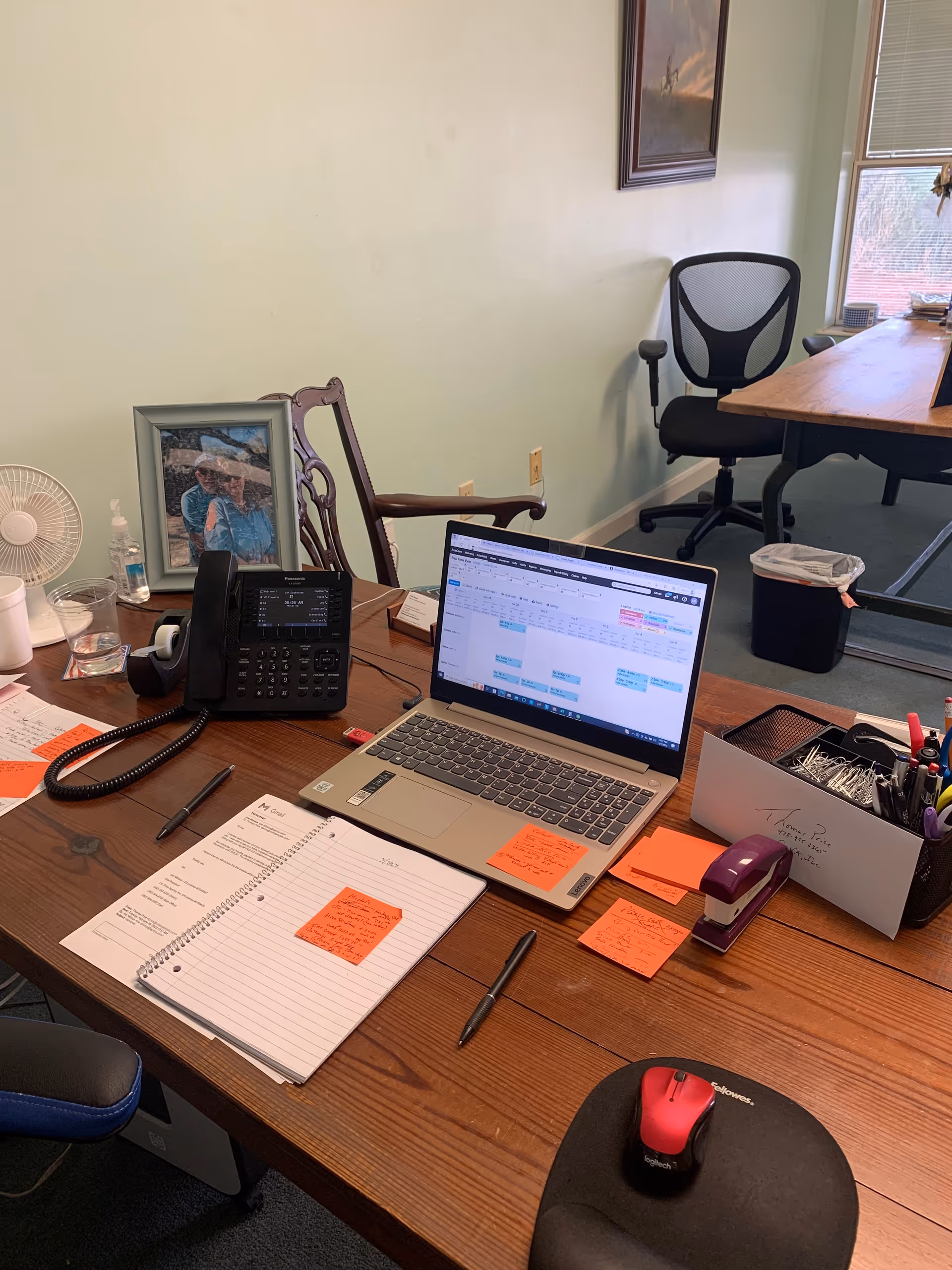 An office desk with a laptop, telephone, notes, a framed photo, and a window view.