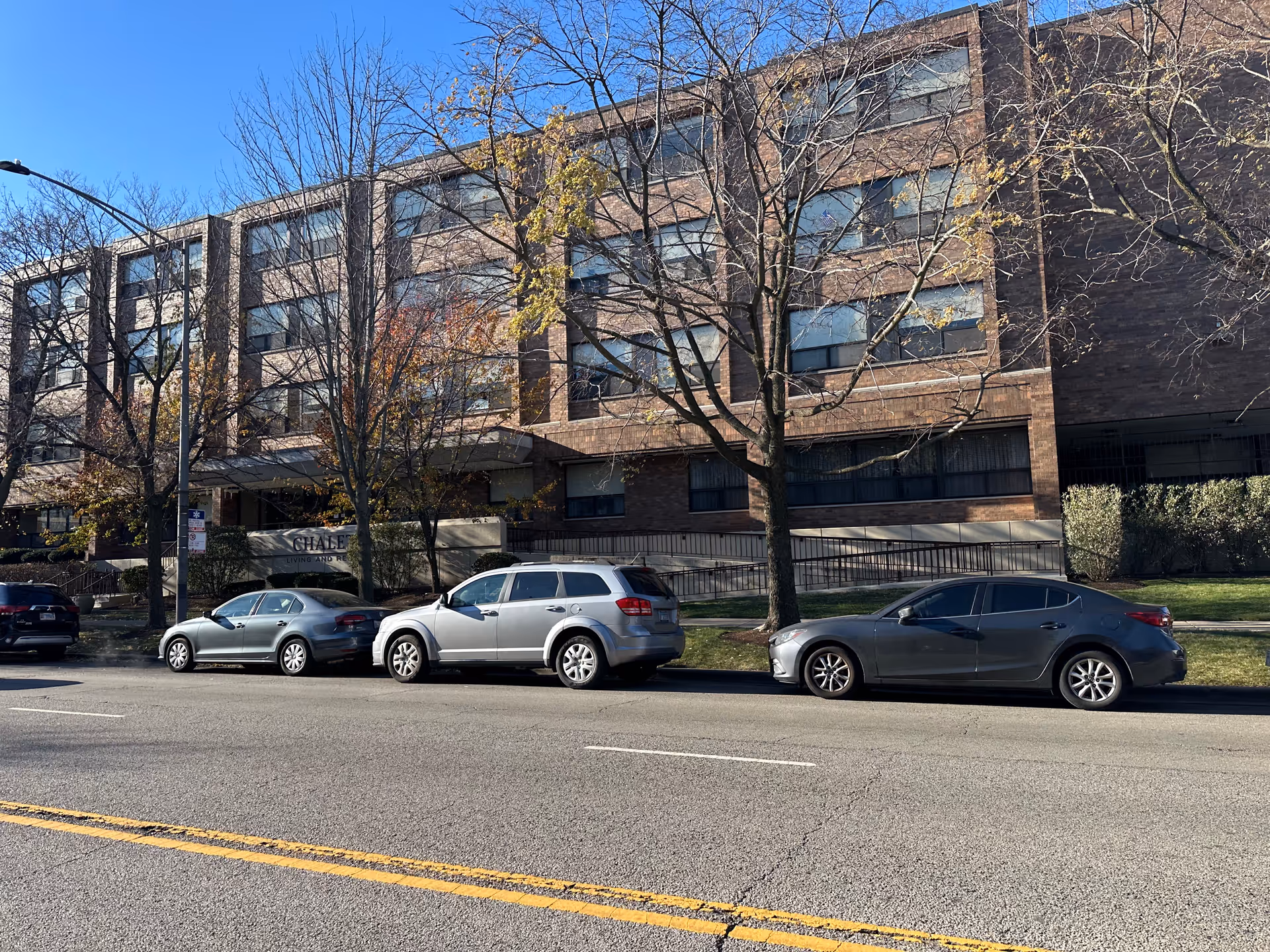 Exterior view of a multi-story brick building with several windows, trees with sparse autumn leaves, and three cars parked along the street in front. A sign near the entrance reads 'Chalet Living And Rehab Center'.