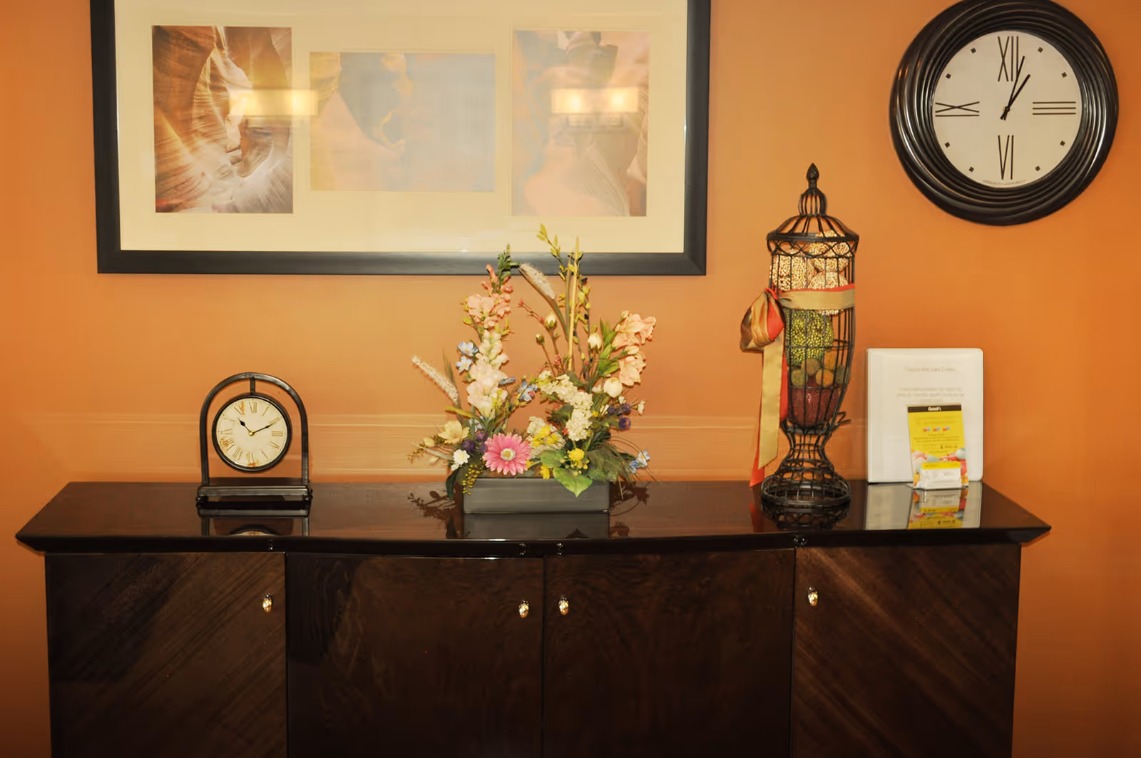 A dark wooden cabinet against an orange wall with a framed picture above it. On the cabinet are a small clock, a floral arrangement in a rectangular vase, a decorative wire container with a ribbon, and some documents including a binder and a small yellow card.