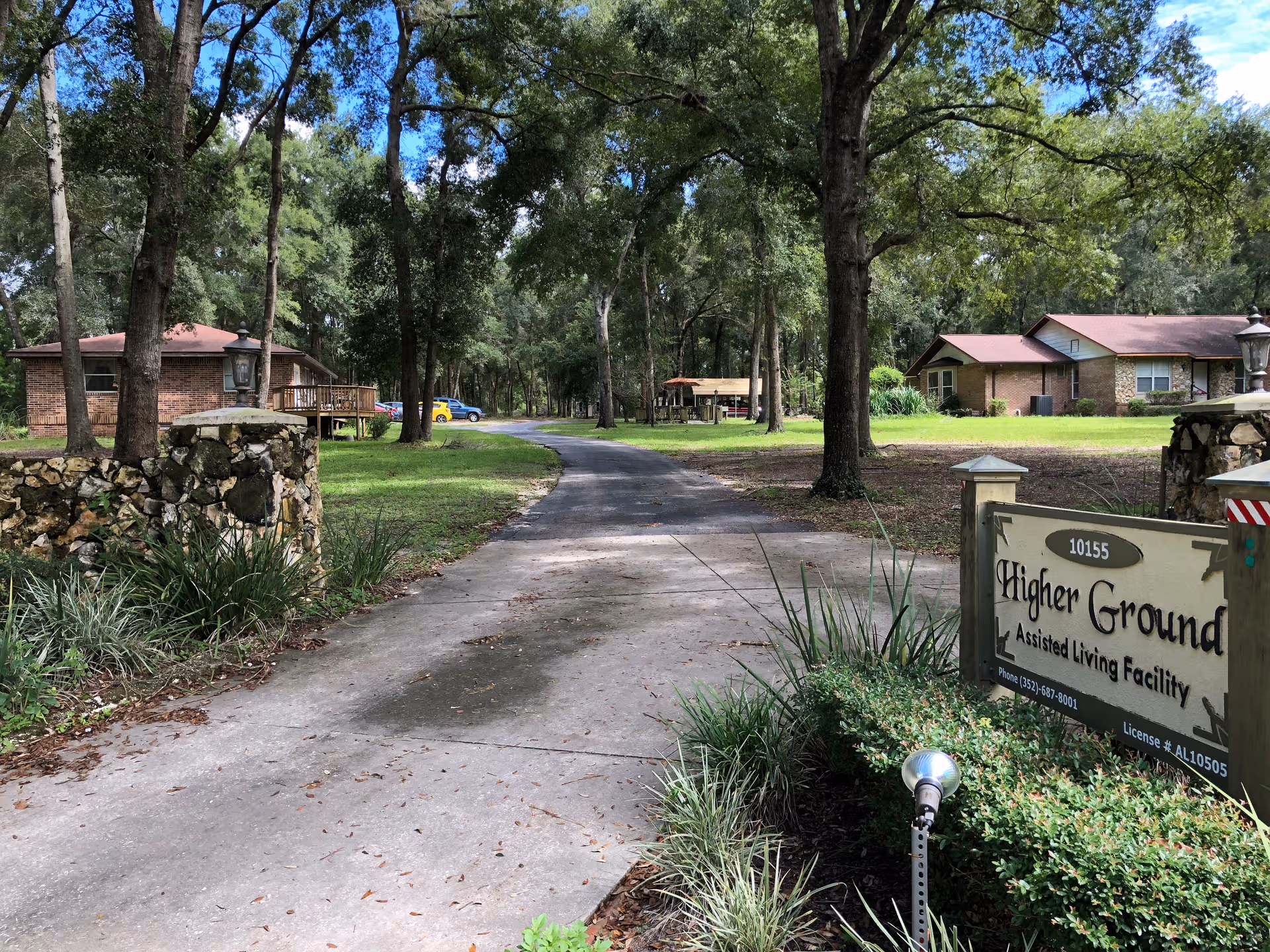 Tree-lined driveway to Higher Ground Assisted Living with a welcoming sign and brick buildings.