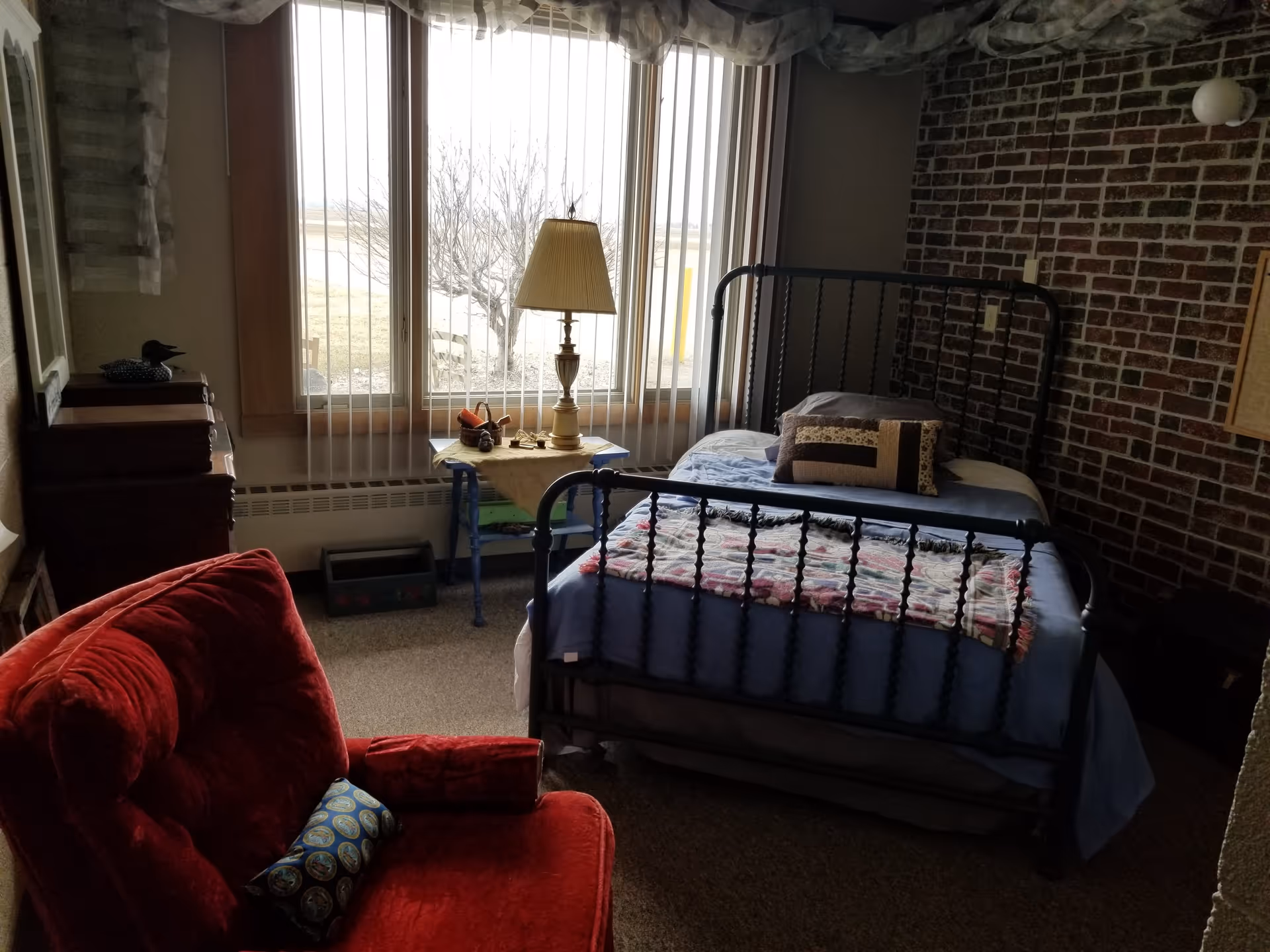 Small bedroom with a metal-framed single bed, red upholstered chair, lamp by a large window with vertical blinds, and a brick accent wall.