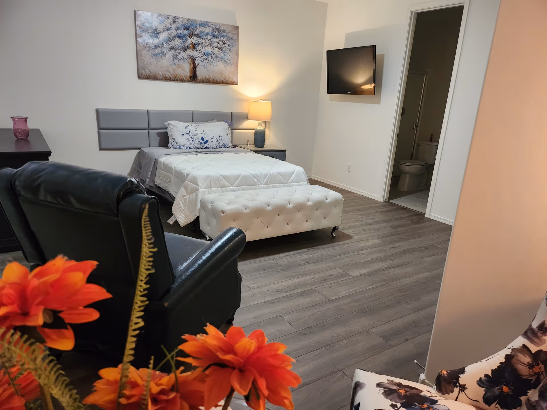 Well-lit senior living bedroom with a bed, seating area, TV, and adjoining bathroom.