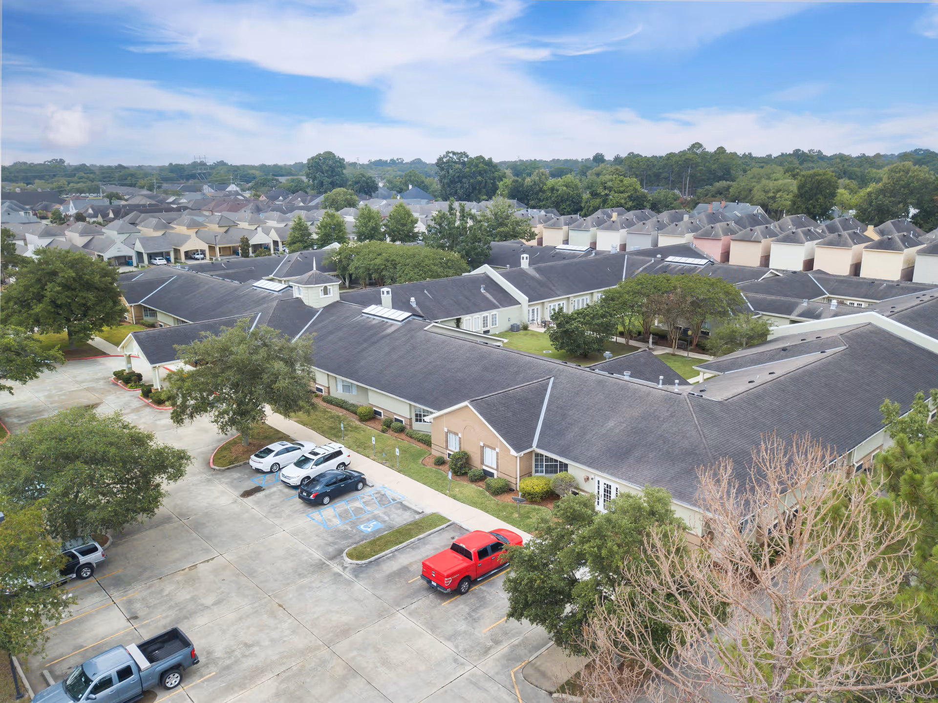 Aerial view of Brookdale Lafayette senior living facility showing multiple connected buildings with dark roofs, surrounded by trees and greenery. There is a parking lot with several cars parked, including a red truck. In the background, there are residential houses and a partly cloudy sky.