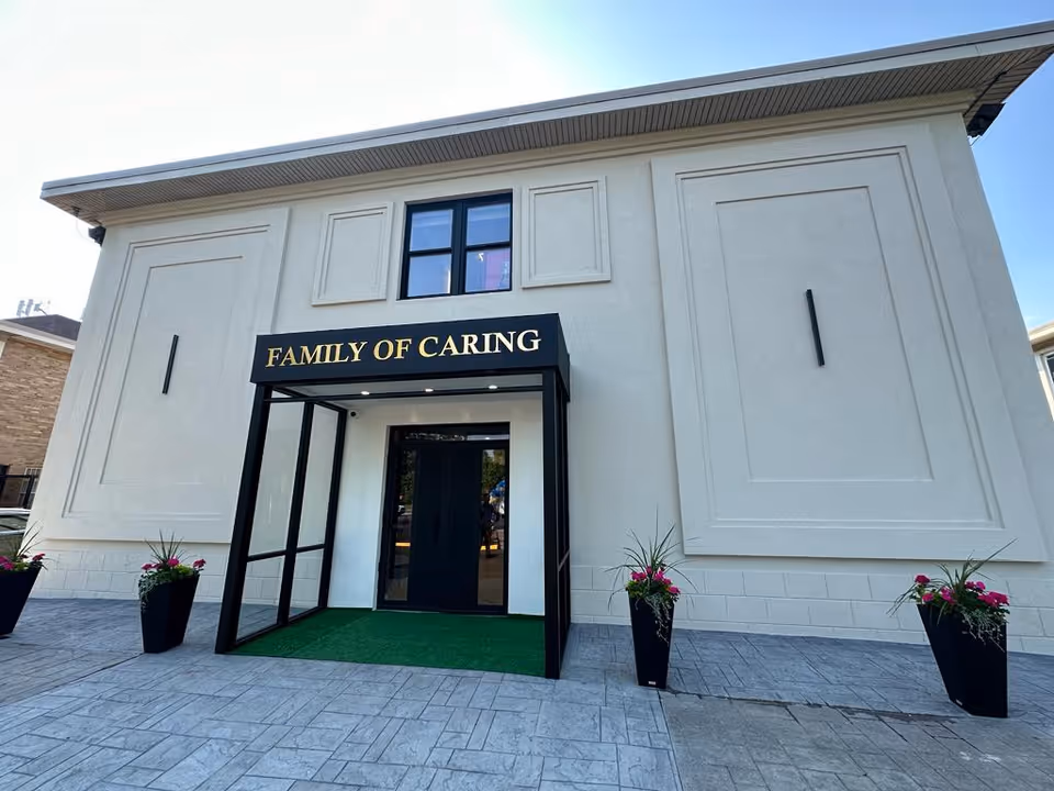 Exterior entrance of a light building with a black 'Family of Caring' sign and green mat.
