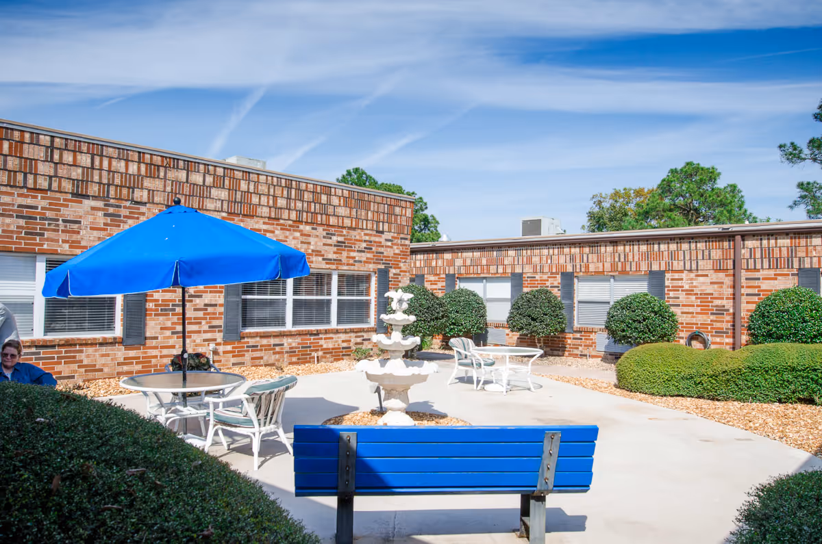 Sunlit brick courtyard with a blue bench, patio tables and umbrella, a tiered fountain, and trimmed shrubs in front of a single-story building.