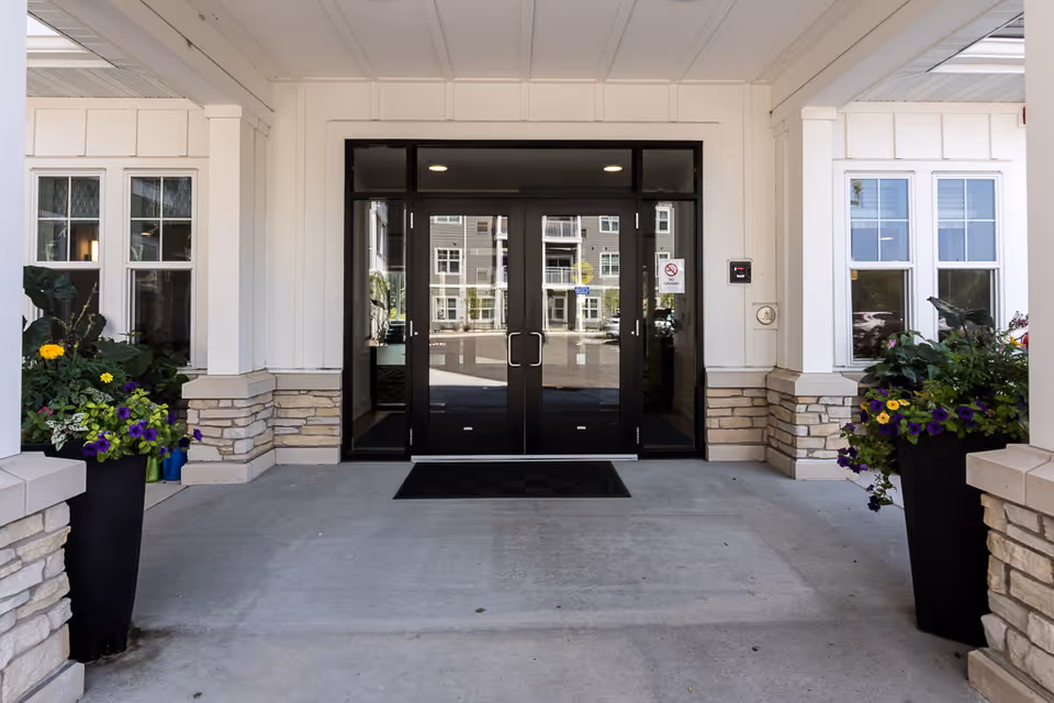 Entrance to a senior living facility with double glass doors framed in black, flanked by white walls with stone accents at the base. Two large black planters with colorful flowers are placed on either side of the entrance. The reflection in the glass doors shows a building and parked cars.