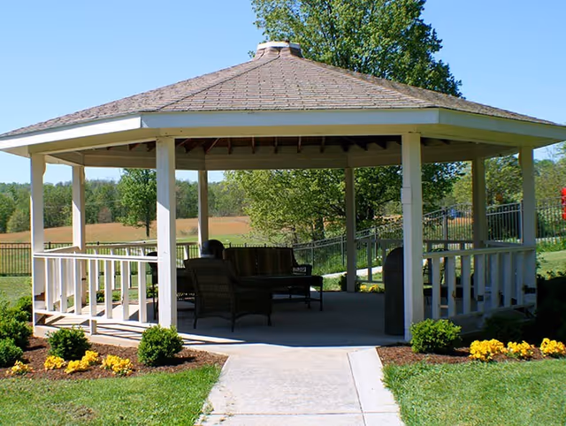 A white wooden gazebo with a shingled roof situated in a grassy outdoor area. Inside the gazebo, there are several dark wicker chairs and a bench arranged around a small table. The surrounding area features green grass, small bushes, and yellow flowers, with trees and a fence in the background under a clear blue sky.