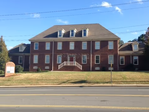 Brick senior living facility with central entrance, steps, and green lawn along a roadside.
