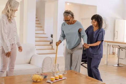 Elderly resident uses a cane with caregiver assistance in a living room, joined by another resident.