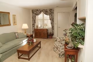 A cozy living room with a green sofa, floral patterned armchairs, a wooden coffee table with a floral centerpiece, a side table with a lamp, a wooden cabinet under a window with floral curtains, and various plants and decorative items.