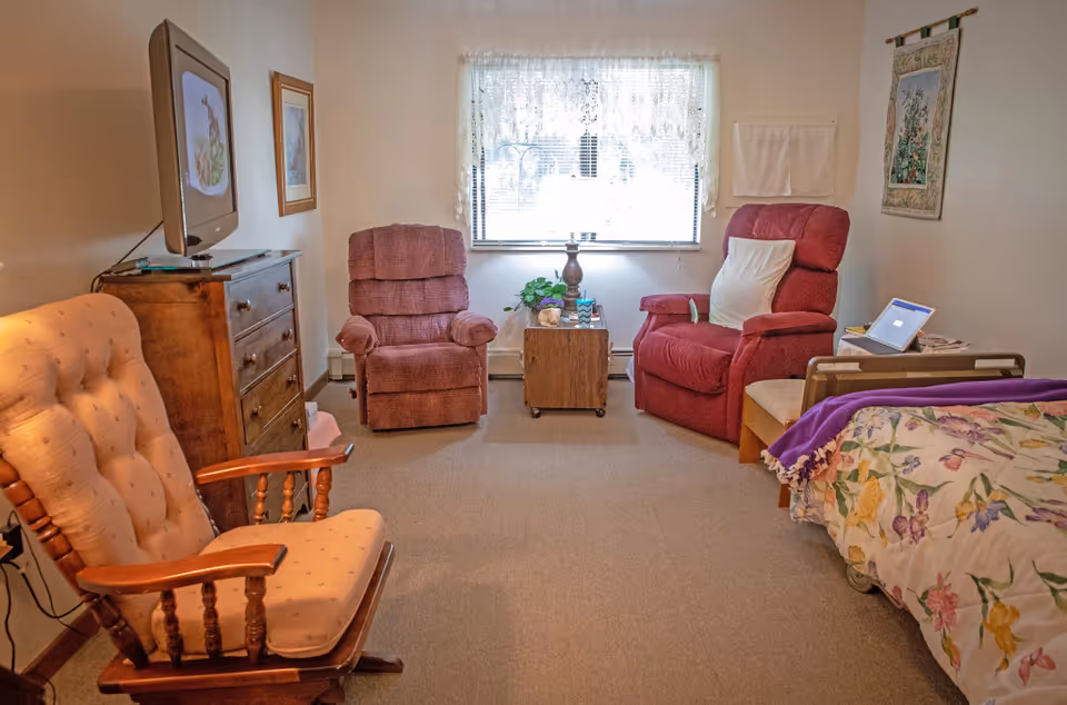 Cozy senior living bedroom featuring a bed, armchairs, dresser, TV, and window.