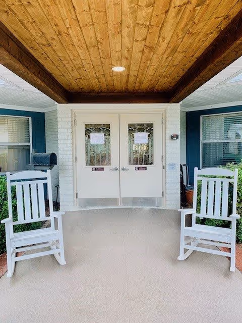 Covered entrance area of a building with two white rocking chairs on either side of double doors. The ceiling above the entrance is wooden with recessed lighting. The building exterior features white brick and blue siding with windows on both sides of the doors.