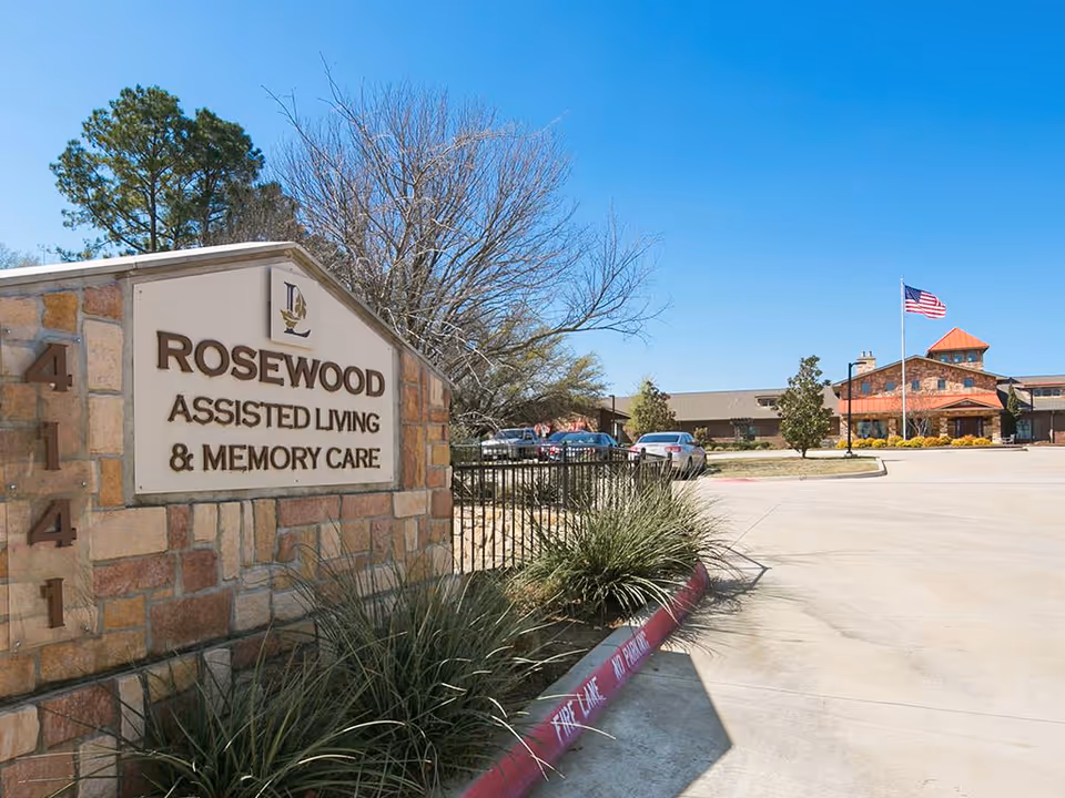 Entrance sign and front driveway of Rosewood Assisted Living & Memory Care with the facility building and American flag in the background.