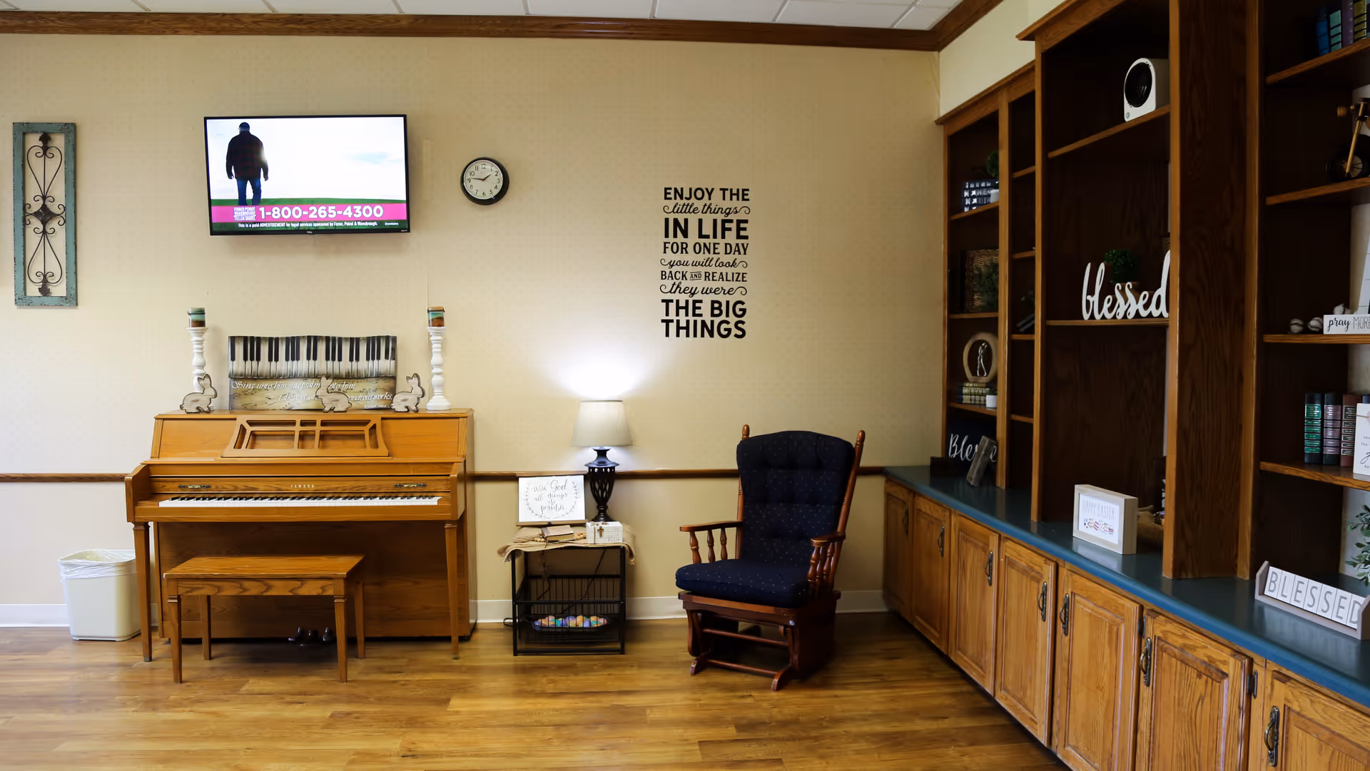 A cozy room with a wooden piano and bench on the left, a small side table with a lamp and books, a cushioned rocking chair, and built-in wooden shelves filled with decorative items and books. A wall clock and a flat-screen TV are mounted on the wall above the piano. The wall features a motivational quote decal.