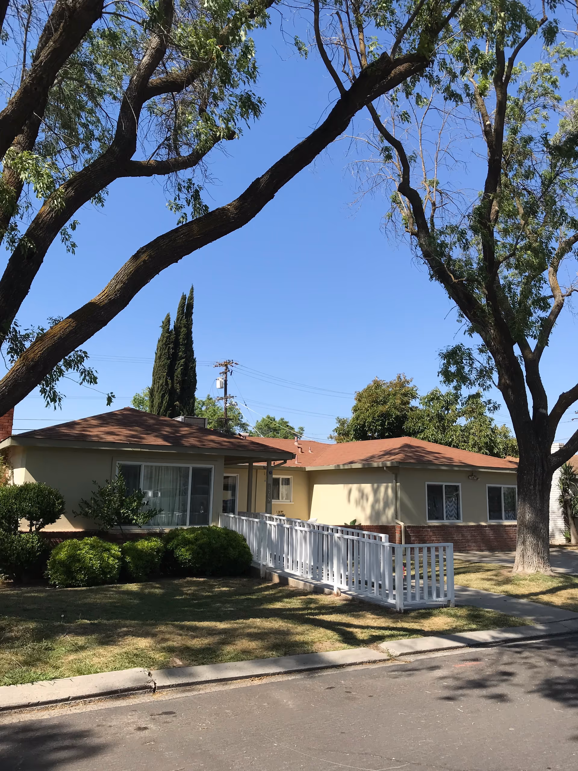 Single-story beige senior living facility with white ramp, entrance, and surrounding trees.
