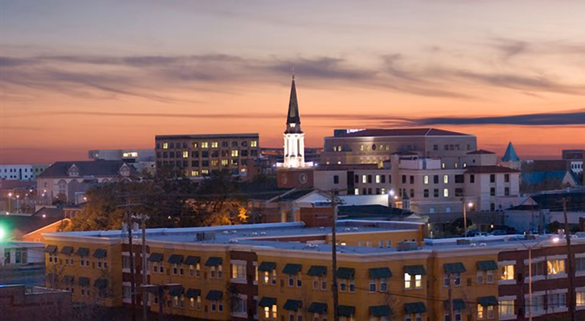 Sunset cityscape showing low-rise buildings and a lit church steeple against a colorful sky.