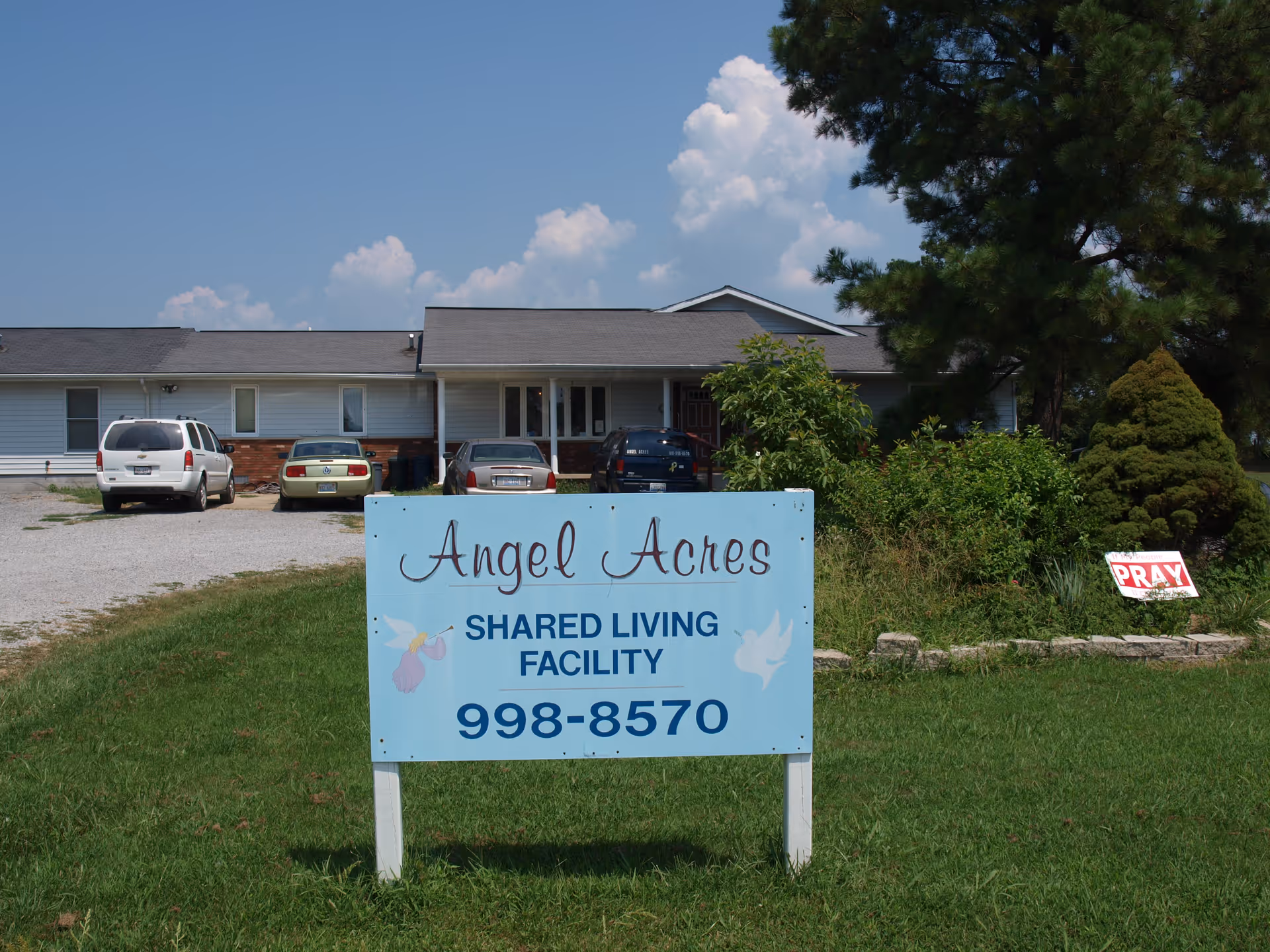 A single-story building with a gravel parking area in front containing several parked cars. In the foreground, there is a blue sign that reads 'Angel Acres SHARED LIVING FACILITY 998-8570' with illustrations of an angel and a dove. The building is surrounded by grass, bushes, and trees under a partly cloudy sky.