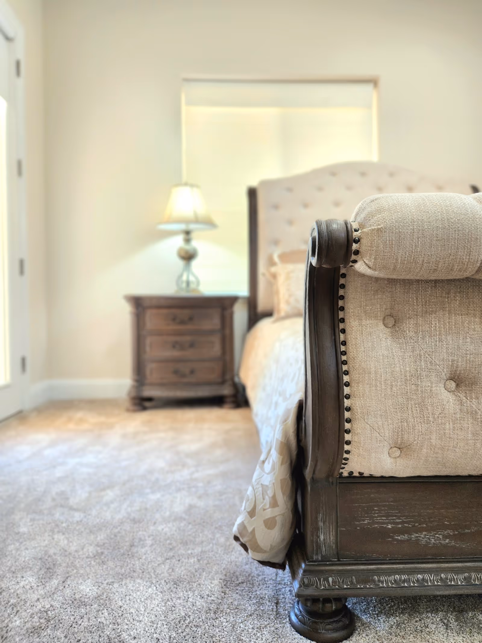 A cozy bedroom features an upholstered bed, bedside table with lamp, and soft carpeting.