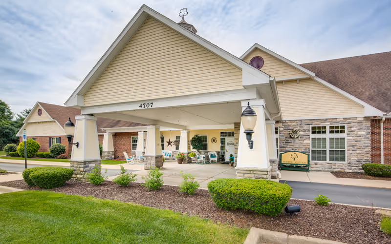 Entrance of a senior living facility named Bickford Of Portage featuring a covered drop-off area with white pillars and stone bases, outdoor seating with chairs and potted plants, well-maintained landscaping with green bushes and grass, and a sign displaying the facility's name on the right side of the building.