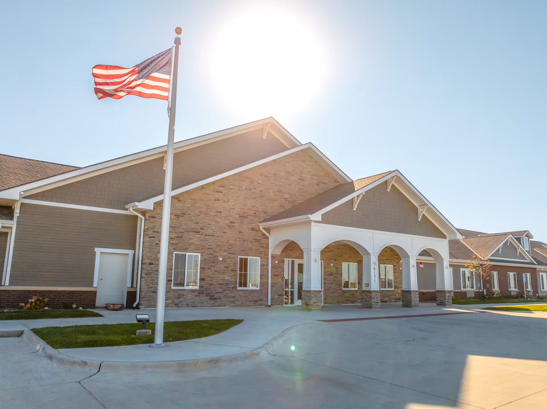 Senior living facility exterior with brick and stone façade, arched entry, flagpole, sunny sky.