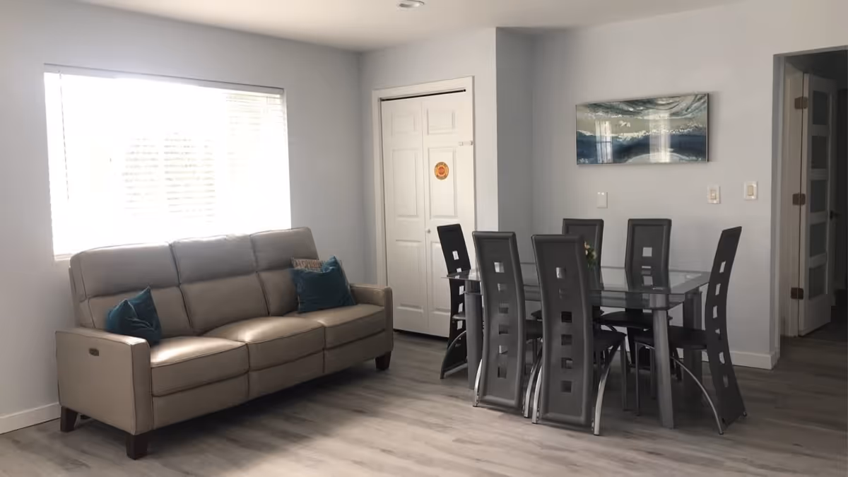 A modern living and dining area featuring a beige leather sofa with teal cushions near a large window with blinds. Adjacent to the sofa is a glass dining table with six black high-back chairs. The room has light gray walls, a white closet door, and a piece of abstract artwork hanging above the dining table. The floor is covered with light wood laminate.