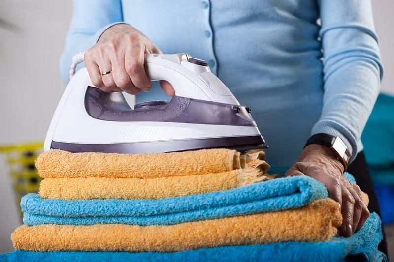 Care worker irons folded towels indoors in a facility.