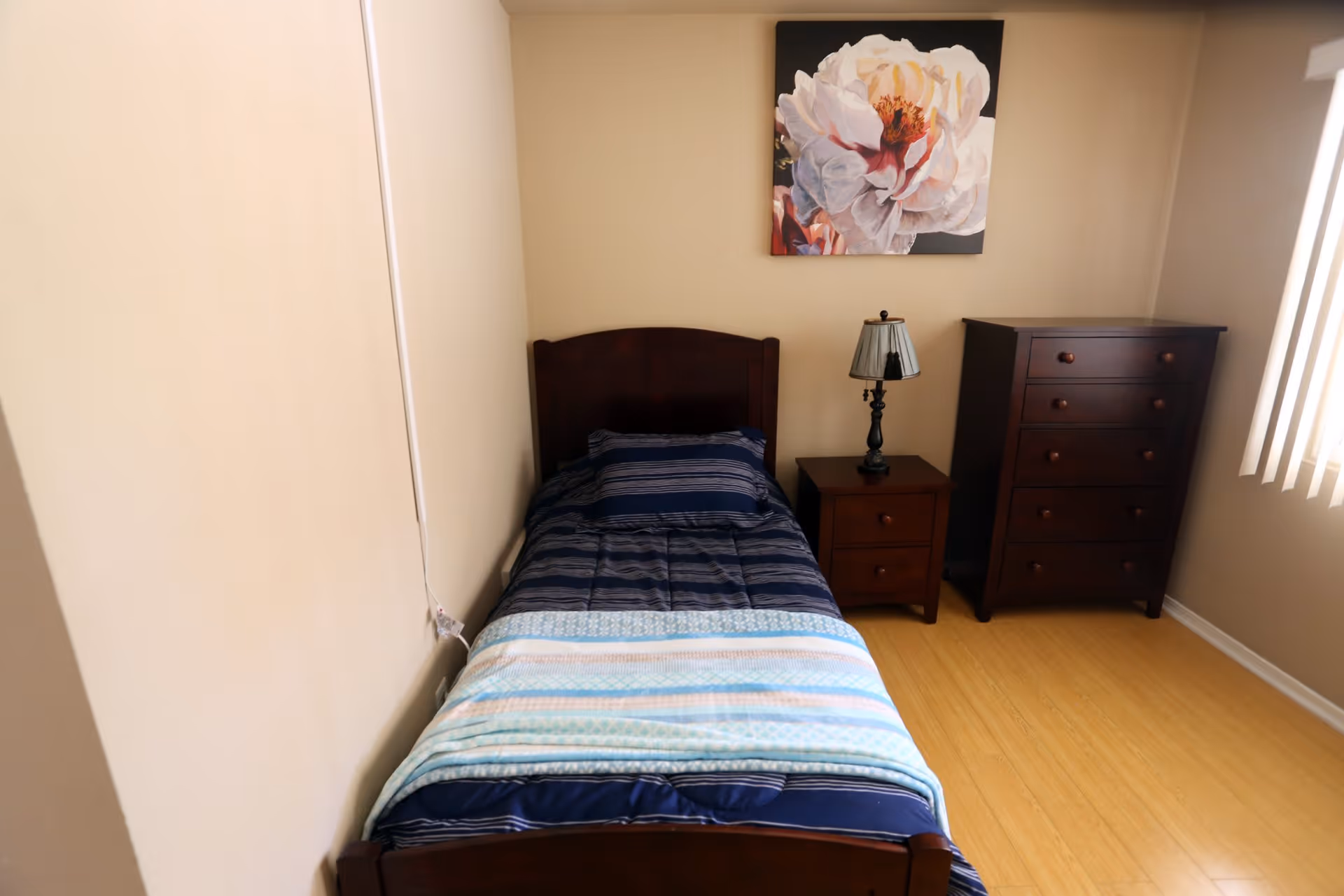 A small bedroom with a single dark wooden bed made with a blue striped comforter and a light blue blanket at the foot. Next to the bed is a matching dark wooden nightstand with a decorative lamp. A dark wooden chest of drawers is positioned against the wall near a window with vertical blinds. Above the bed hangs a large floral painting with white and pink flowers. The room has light beige walls and a light wooden floor.