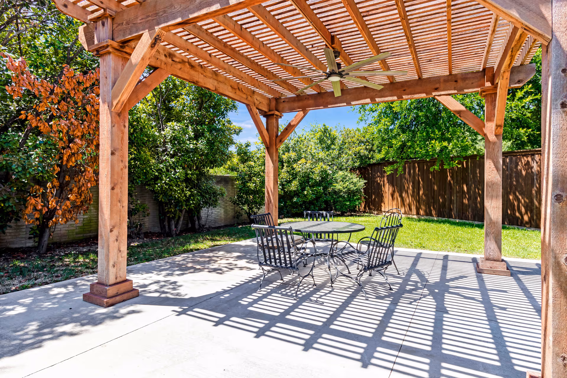 Outdoor patio area with a wooden pergola casting shadows on the concrete floor. Under the pergola is a round metal table with four matching chairs. Surrounding the patio are green trees and bushes, with a wooden fence in the background.