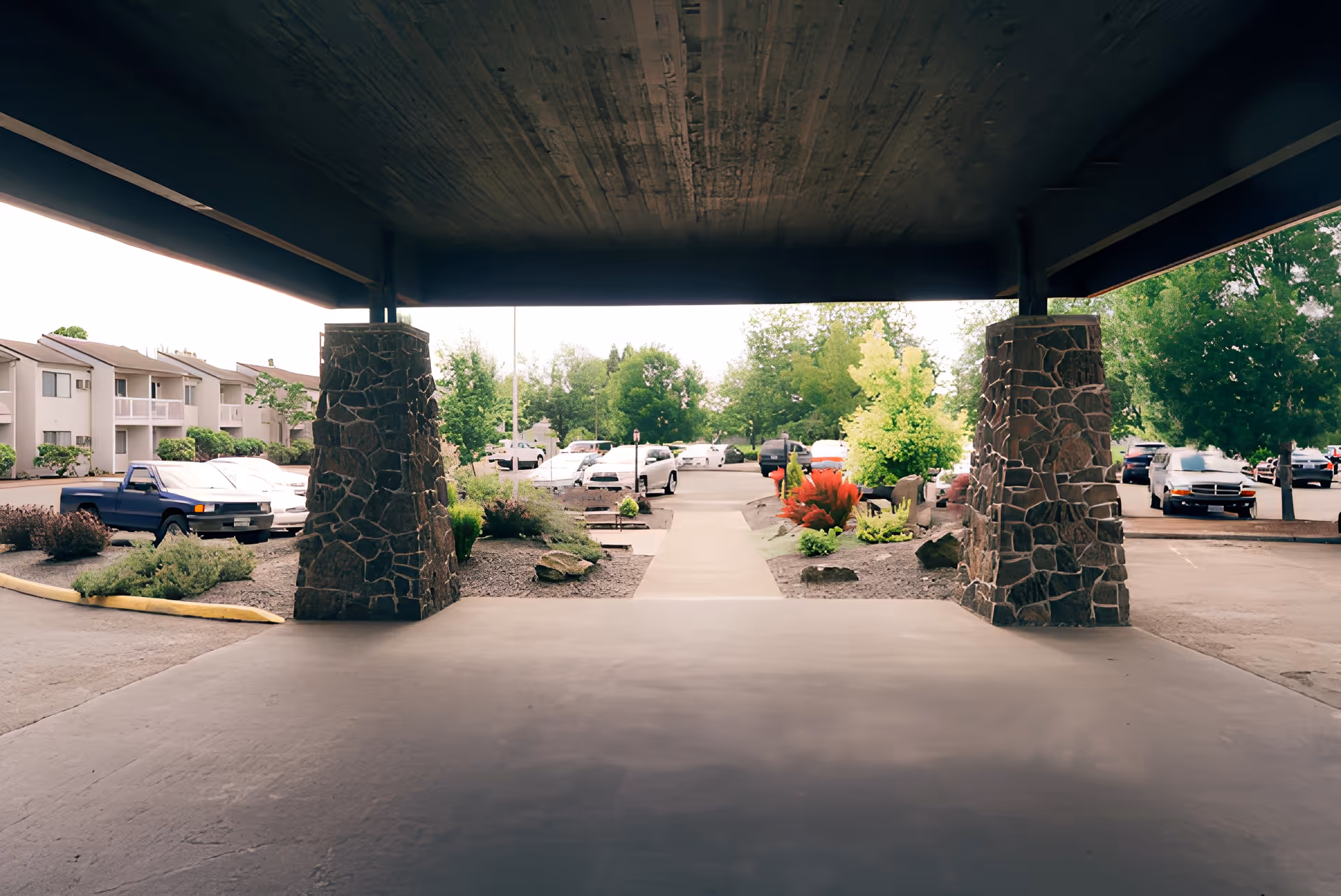 Covered porte-cochere with stone pillars opening onto a landscaped walkway, parking lot, and apartment-style buildings in the background.