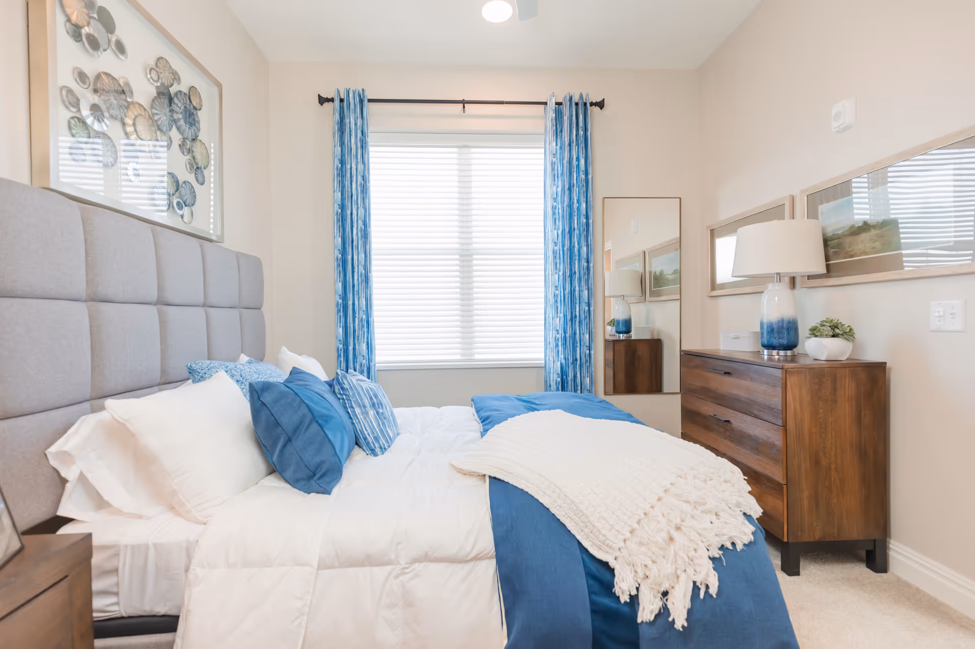 A neatly made bed with white and blue bedding and multiple pillows in a bedroom. The room features a large window with blue curtains, a wooden dresser with a lamp and small plant, a full-length mirror, and framed artwork on the walls.