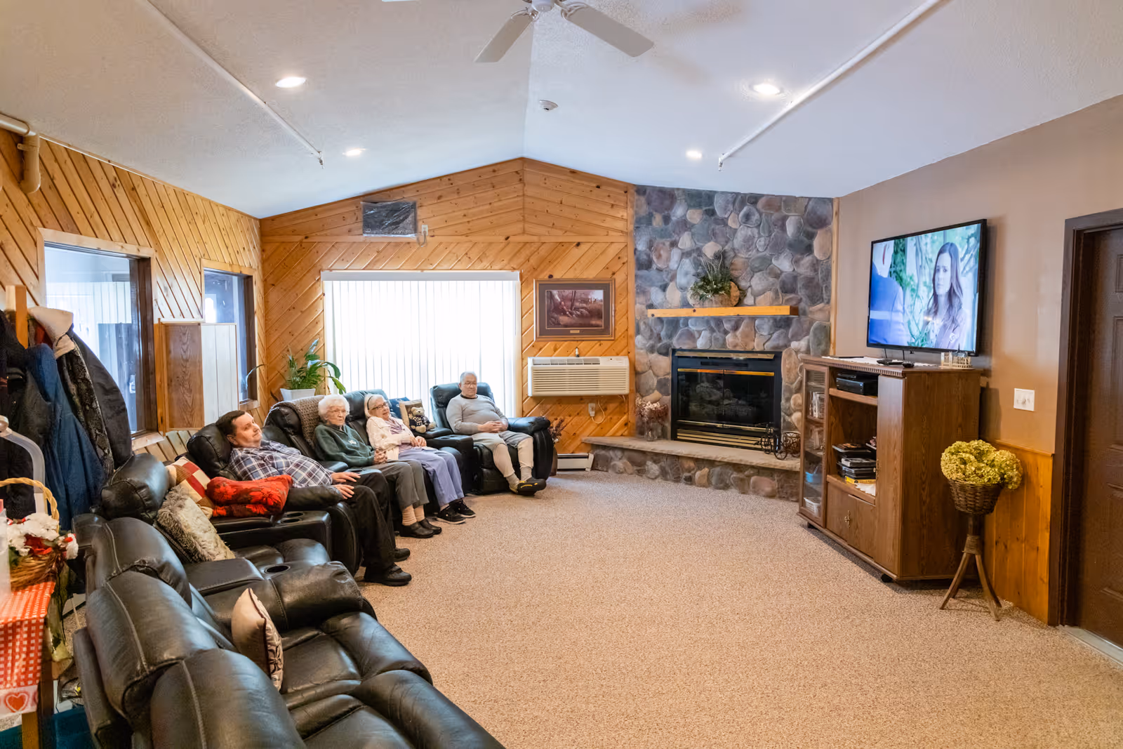 A cozy living room in a senior living facility with wood-paneled walls and a stone fireplace. Four elderly people are seated comfortably on recliners along the left wall, watching a television mounted on the right wall. The room is carpeted and has a ceiling fan, with a cabinet and a basket of flowers near the TV.