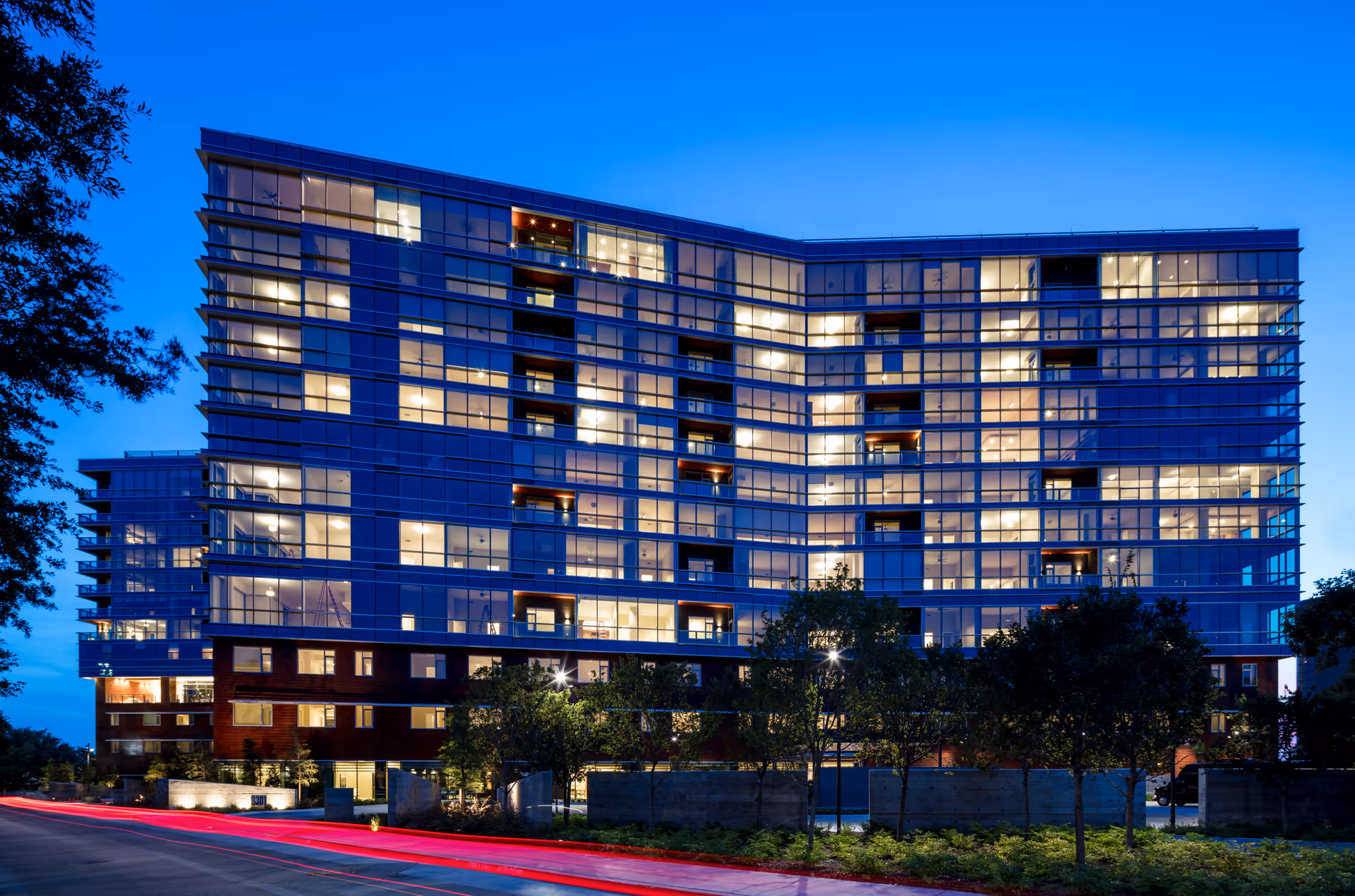 Exterior view of a modern multi-story building at dusk with many illuminated windows and some trees in front.