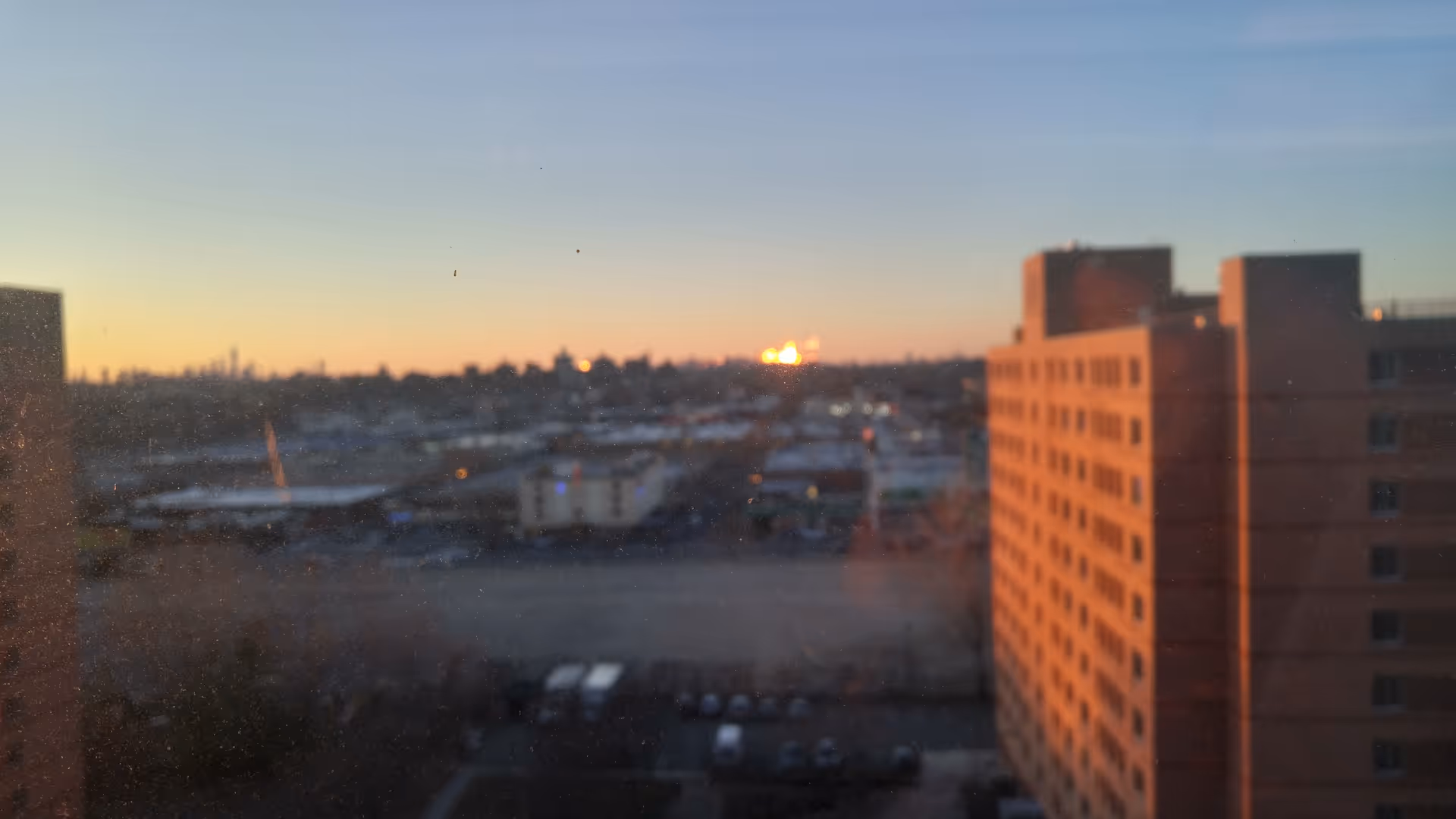 Sunset over a city skyline seen from a window beside a tall brick building.
