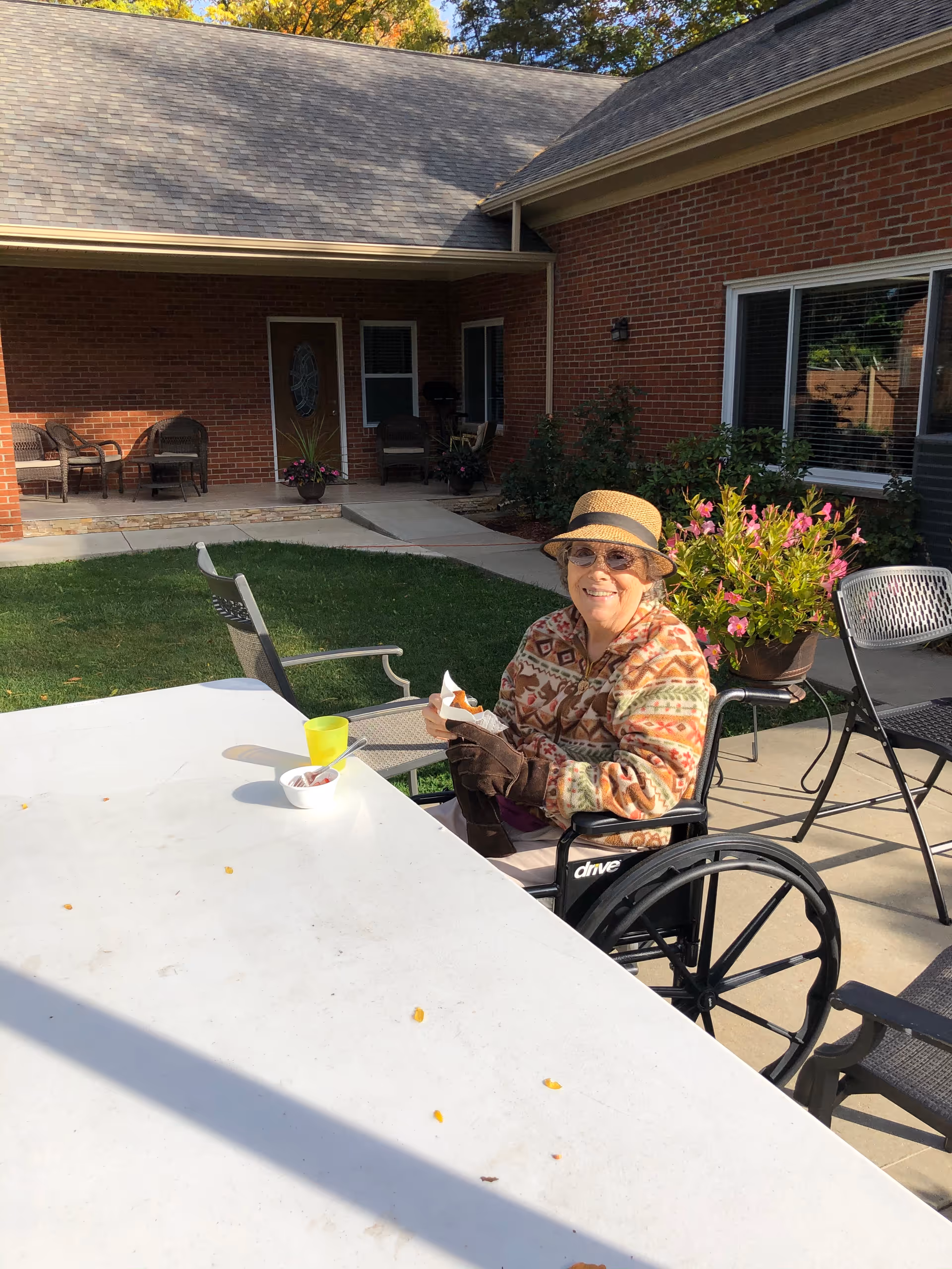 An elderly woman wearing a patterned jacket, gloves, sunglasses, and a hat is sitting in a wheelchair outdoors at a white table. She is smiling and holding a napkin. There is a yellow cup and a bowl on the table. Behind her is a brick building with windows, a door, patio chairs, and potted plants with flowers. The scene is sunny with green grass and trees in the background.