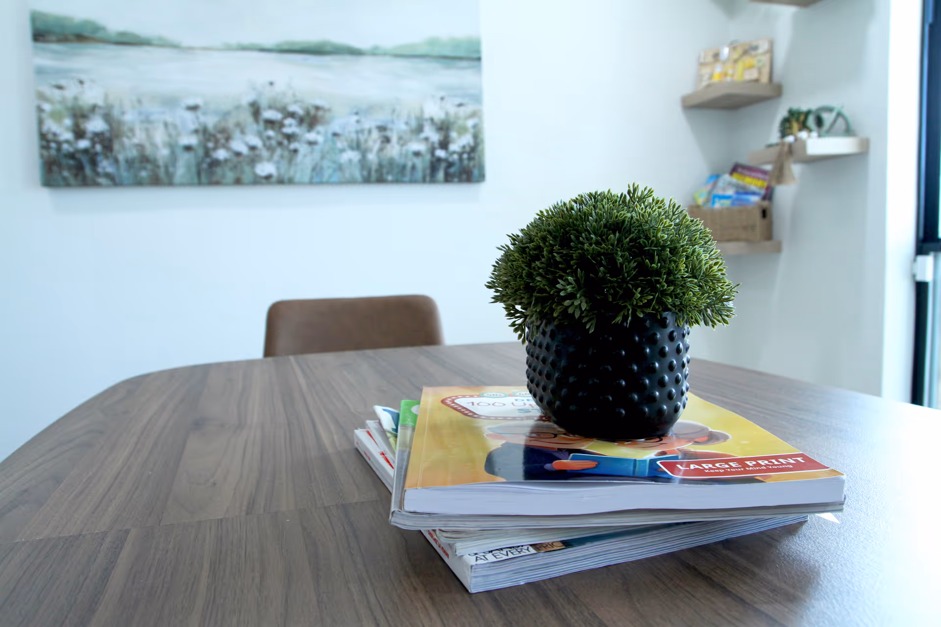 Wooden table with a black textured plant pot on magazines inside a bright lounge area.