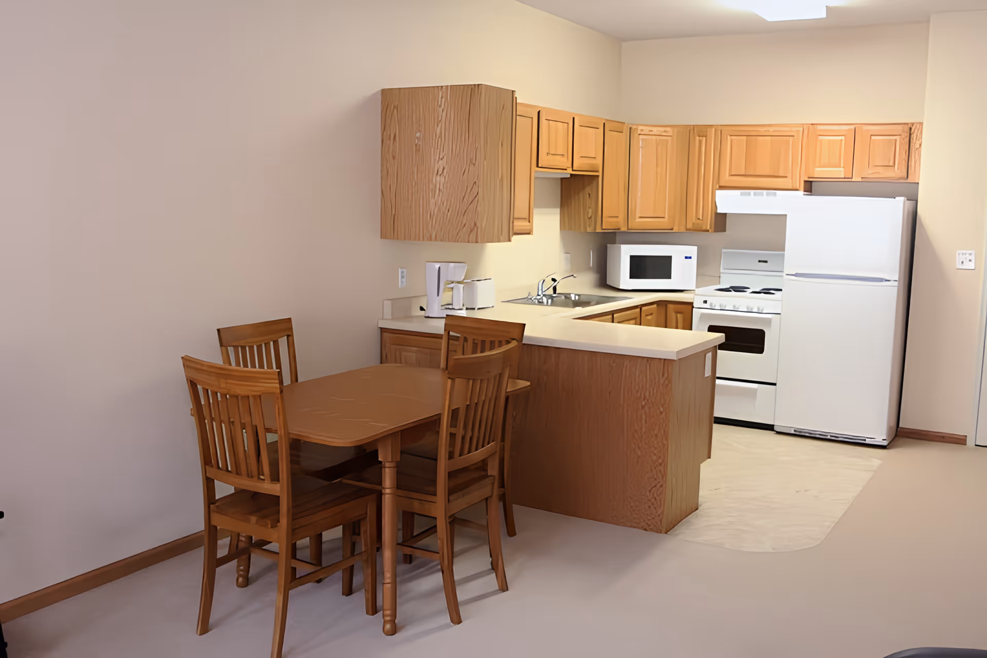 A small kitchen and dining area in an assisted living facility. The kitchen features wooden cabinets, a white refrigerator, stove, microwave, and a coffee maker on the counter. Adjacent to the kitchen is a wooden dining table with four matching chairs. The walls are painted a light beige color.