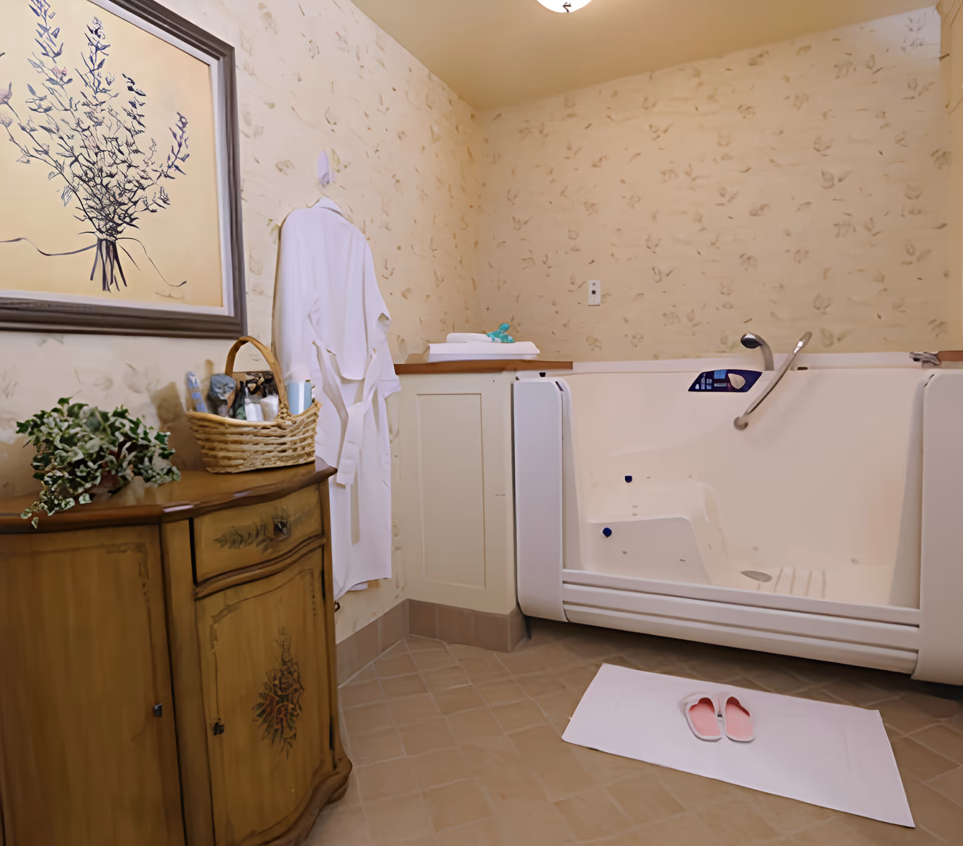 A bathroom with a walk-in bathtub featuring a side door and grab bars. A white bathrobe hangs on the wall next to a wooden cabinet with a basket and a potted plant on top. A pair of pink slippers is placed on a white bath mat on the tiled floor. The walls have a light beige textured pattern, and a framed artwork of a plant hangs above the cabinet.